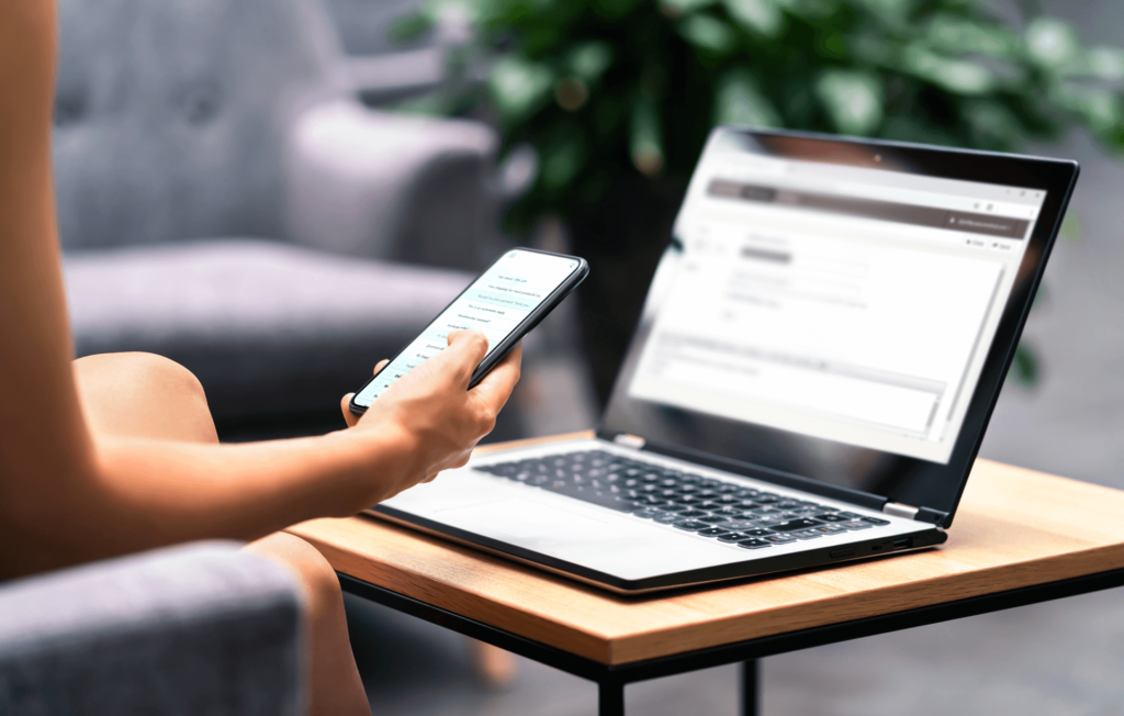 Person uses laptop on wooden table while holding a smartphone